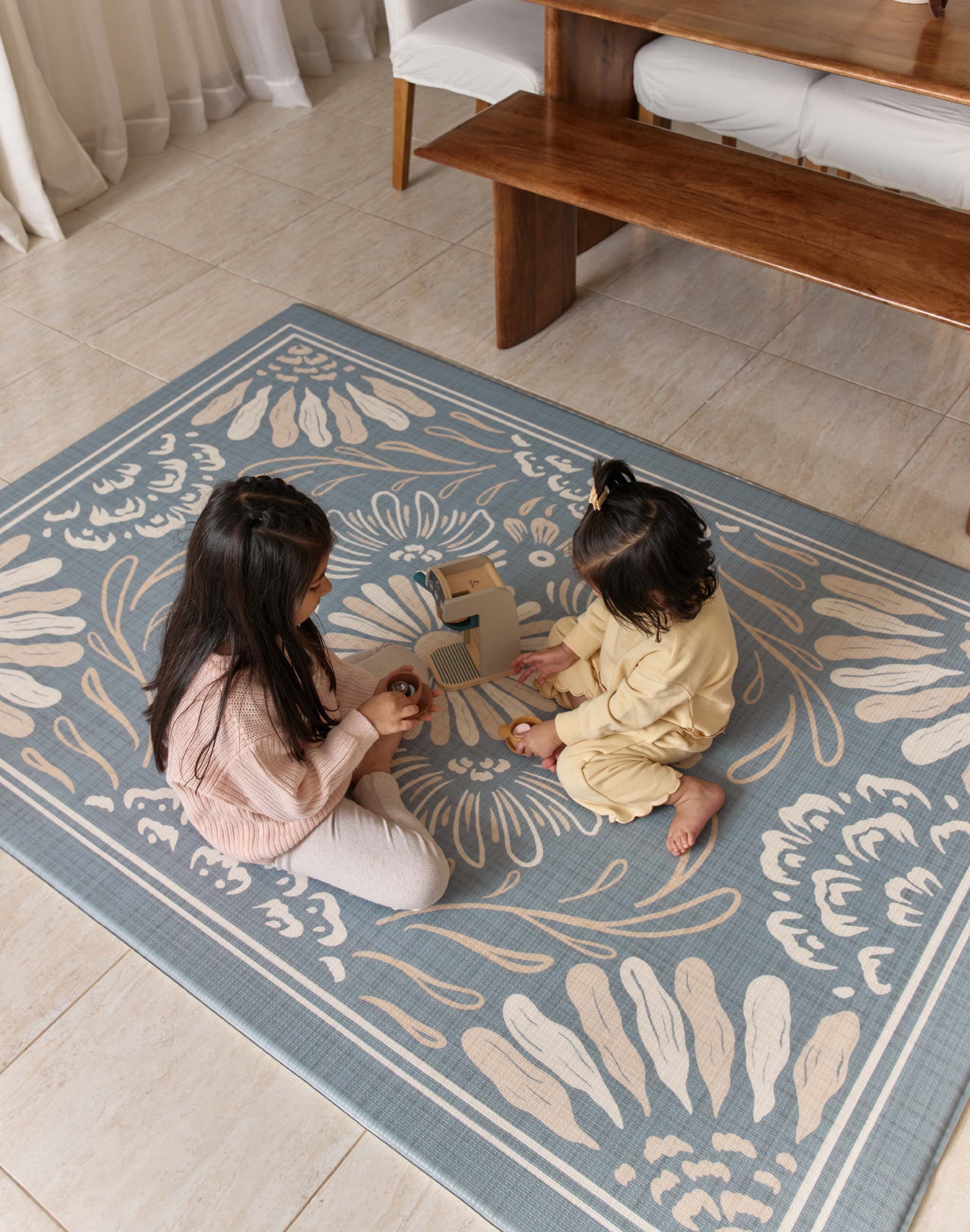 Two children sitting on a decorative rug in a room with a table and chairs.